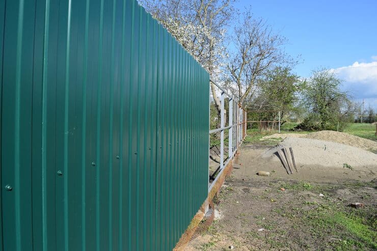 A green metal fence is partially installed on a grassy area, with construction materials and a pile of sand nearby. Leafy trees are in the background under a blue sky with scattered clouds.