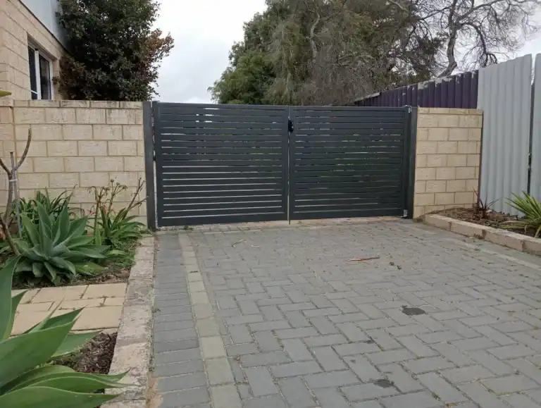 A closed black metal gate with horizontal slats set in a beige brick wall, opening onto a driveway paved with grey bricks. Green plants grow along the edges, and trees are visible behind the gate.