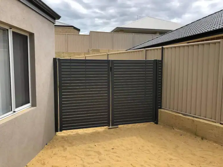 A small back garden area with sandy ground, surrounded by beige metal panel fencing; a dark grey slatted metal gate is fitted between two sections of the fence, next to a house with a window.