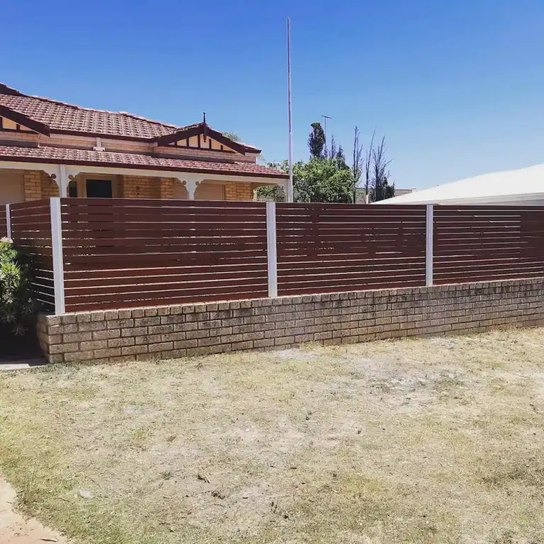 A brick house with a red tiled roof is behind a low brick wall topped with a modern horizontal slat fence and white posts, on a sunny day with a blue sky and dry grass in the foreground.